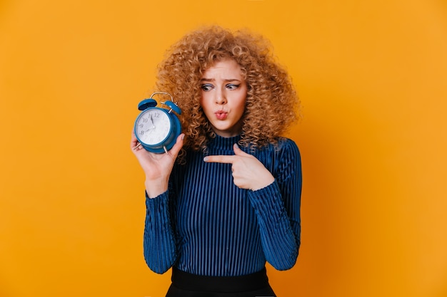 Free photo curly girl in blue striped sweater looks at alarm clock with suspicion and points finger at it.