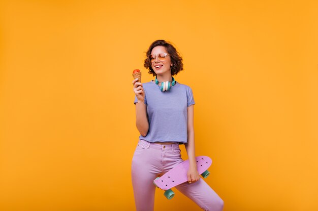 Кудрявая девушка в очках со скейтбордом, стоя в позе уверенно. Внутреннее фото красивой женщины, едящей мороженое.