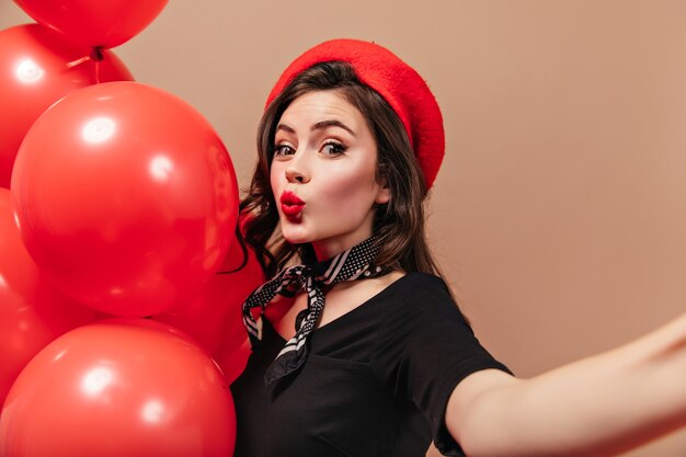 Curly girl in red beret and silk scarf whistles, holds balloons and makes selfie.