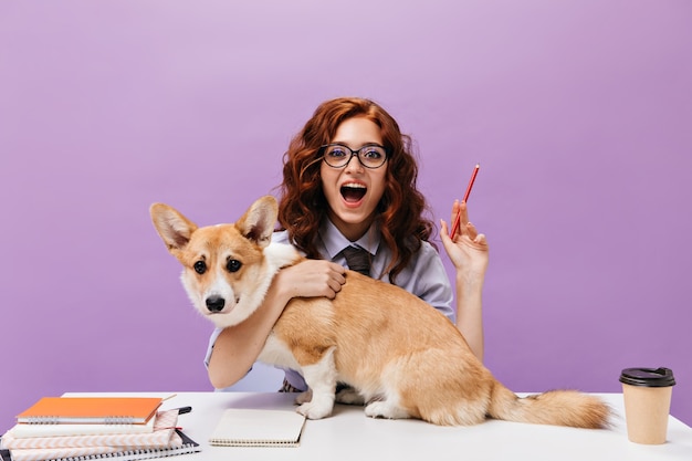 シャツと眼鏡の巻き毛の女の子は犬を抱きしめ、鉛筆を保持します
