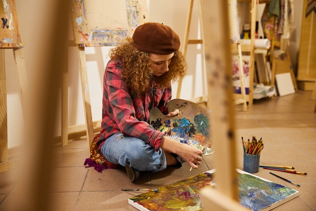 La ragazza riccia si siede su un pavimento, mescolando oli e disegna un dipinto