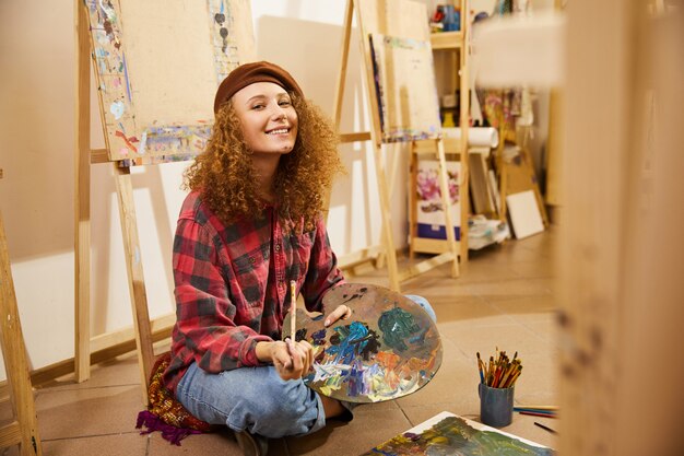 Curly girl sits on a floor, smiling and holds a palette with oils