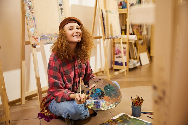 Curly girl sits on a floor, smiling and holds a palette with oils