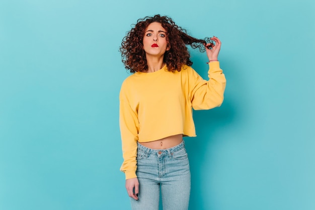 Curly girl in yellow sweatshirt and denim pants looks at camera with funny face and plays hair on blue isolated background