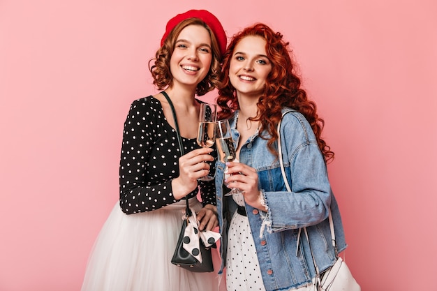 Free photo curly girls clinking wineglasses and laughing. refined friends drinking champagne on pink background.