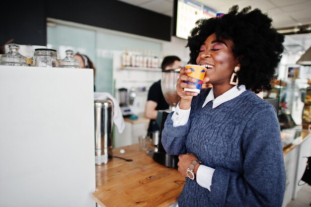 巻き毛のアフリカ系アメリカ人女性は、お茶やコーヒーと一緒に屋内カフェでポーズをとったセーターを着ています