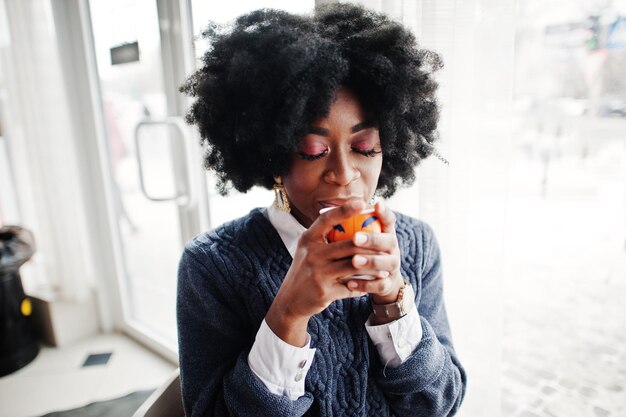 Кудрявая африканская американка в свитере позирует в кафе в помещении с чашкой чая или кофе