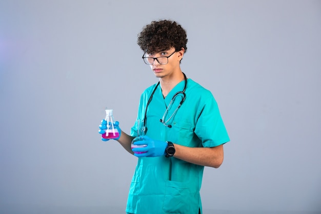 Curly hair boy in medical uniform and hand masks holding chemical flasks on grey background