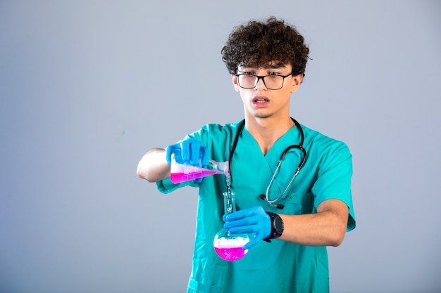 Curly hair boy in medical uniform and hand masks putting pink liquid from one flask to another on grey background. 