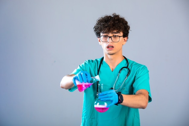 Curly hair boy in medical uniform and hand masks putting pink liquid from one flask to another very carefully.