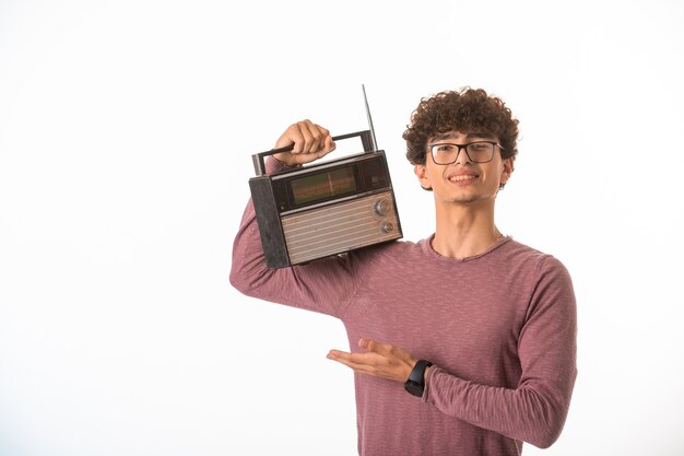 Curly hair boy in optique glasses holding a vintage radio in his shoulders. 