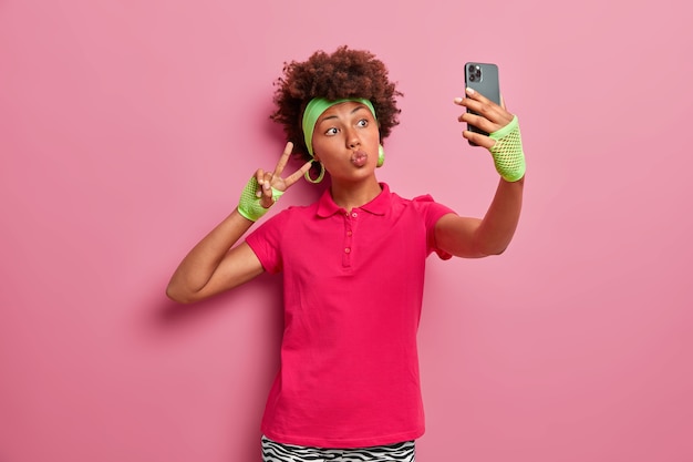 Free photo curly haired active woman in pink t shirt, headband and sport gloves, takes selfie, makes victory gesture, holds mobile phone, being obsessed with social networks poses indoor