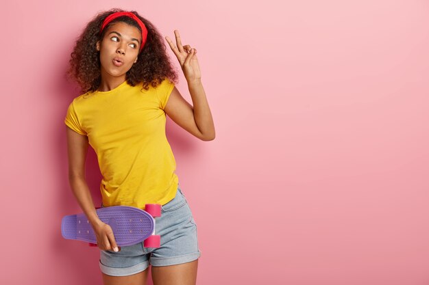 Curly haired beautiful female skater in casual clothes, carries skateboard, makes peace gesture, looks aside