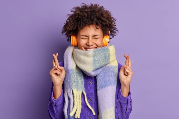 곱슬 머리 민족 소녀는 손가락을 교차하고 행운을 믿고 헤드폰으로 즐거운 노래를 듣고 따뜻한 겨울 스카프를 착용합니다.