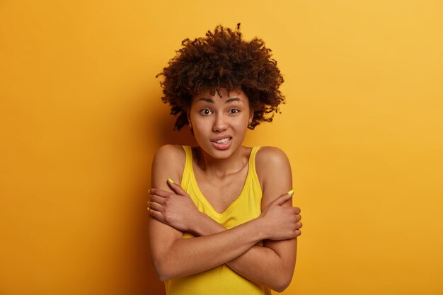 curly haired ethnic woman trembles from cold, crosses arms over body, clatters teeth, feels insecure and scared, wears casual yellow shirt, stands indoor, makes defensive pose, afraids of something