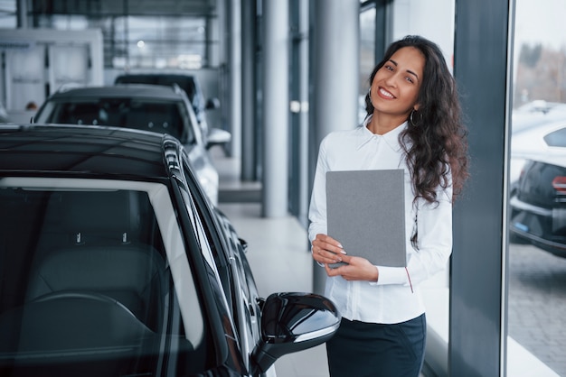 Curly haired female manager stands near the car in automobile salon