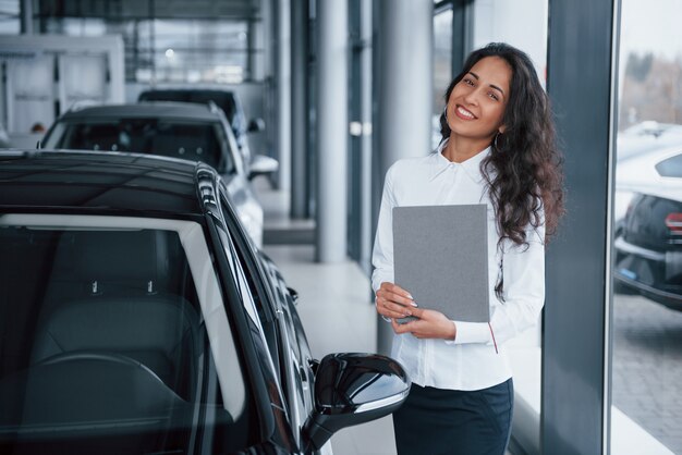 Кудрявая женщина-менеджер стоит возле машины в автомобильном салоне