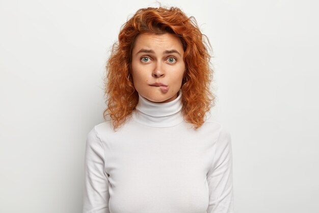 Curly haired ginger female bites lips, has nervous troubled expression, hesitates about something
