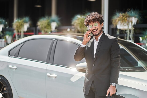 Curly-haired Indian businessman talking on the phone in front of the car.