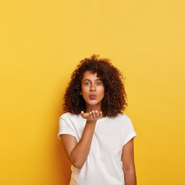 Curly haired lovely Afro girl sends air kiss , holds palm near mouth, dressed in white t shirt, blows passionate mwah, keeps lips folded, models against yellow wall, copy space above