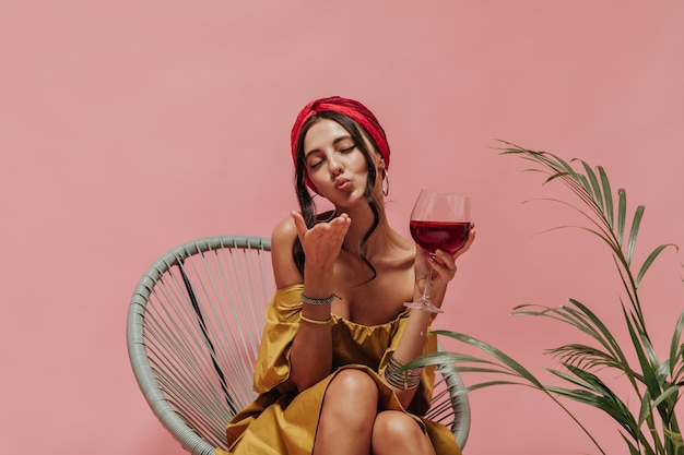 Curly haired pretty lady in red headband and yellow trendy sundress blowing kiss and holding glass on isolated backdrop