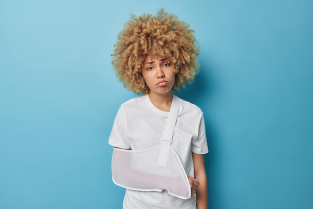 Curly haired upset European woman has problems with health injured or broken arm looks sadly at camera dressed in casual t shirt isolated over blue background Female patient comes to surgeon