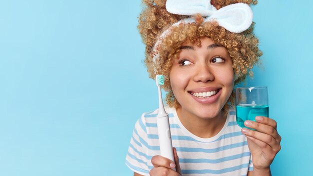 縮れ毛の女性は屋内で歯を磨く歯ブラシを保持し、うがい薬のガラスは彼女の健康の世話をします毎日のルーチンは青い背景で隔離のヘッドバンドカジュアルな縞模様のTシャツを着ています