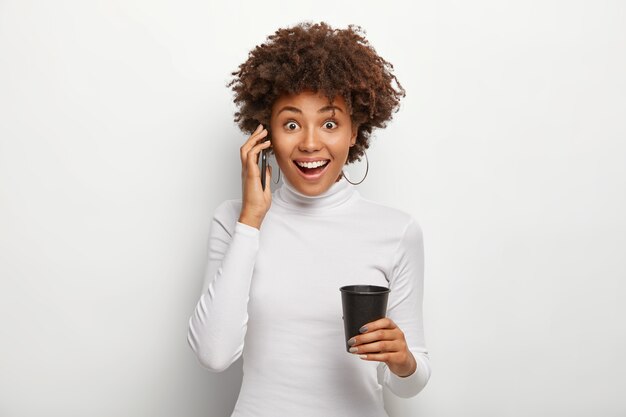 縮れ毛の女性はスマートフォンを介して幸せな会話をし、面白いニュースを聞くのを楽しんで、飲み物の黒い紙コップを保持します