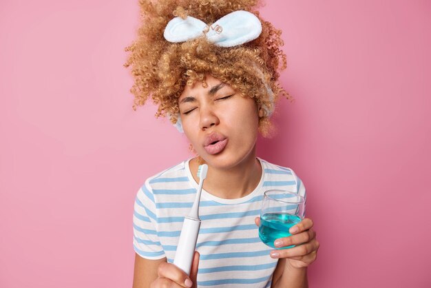 Curly haired woman holds electric brush as if microphone sings song holds glass of mouthwash wears headband and casual striped t shirt isolated over pink background Personal oral hygiene concept