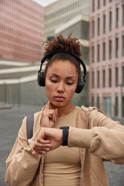 Curly haired woman times pulse looks at smartwatch keeps hand on neck dressed in sportswear listens music via wireless headphones poses against blurred background. Wellness and sport concept