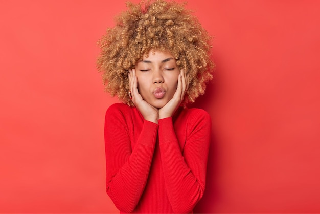 Foto gratuita la giovane donna dai capelli ricci mantiene le labbra arrotondate attende il bacio chiude gli occhi ha un'espressione romantica indossa un maglione casual a maniche lunghe isolato su sfondo rosso vivido. concetto di persone e sentimenti