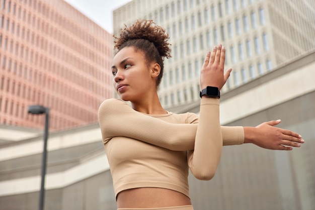 Foto gratuita la giovane donna dai capelli riccia allunga le braccia si riscalda prima dell'allenamento vestita con uno smartwatch ritagliato in posa all'aperto