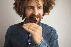 Free photo curly happy man with healthy skin bites homemade artisan chocolate bar, while smiling, isolated on white, wearing jean shirt