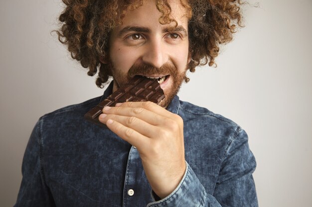 Curly happy man with healthy skin bites homemade artisan chocolate bar, while smiling, isolated on white, wearing jean shirt