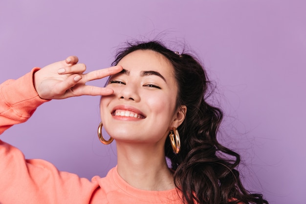 Curly japanese woman showing peace sign. Stunning asian female model in earrings laughing at camera.