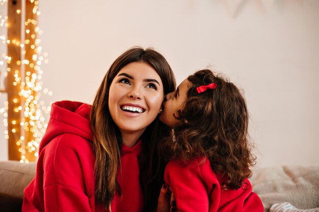 Curly kid kissing mom at home. Young mother enjoying leisure time with daughter.