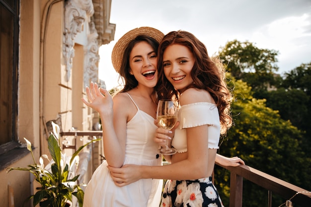 Curly ladies hug and pose with champagne glasses on balcony