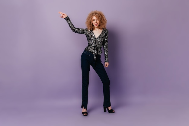 Curly lady in shiny blouse and jeans dancing disco on purple isolated space.