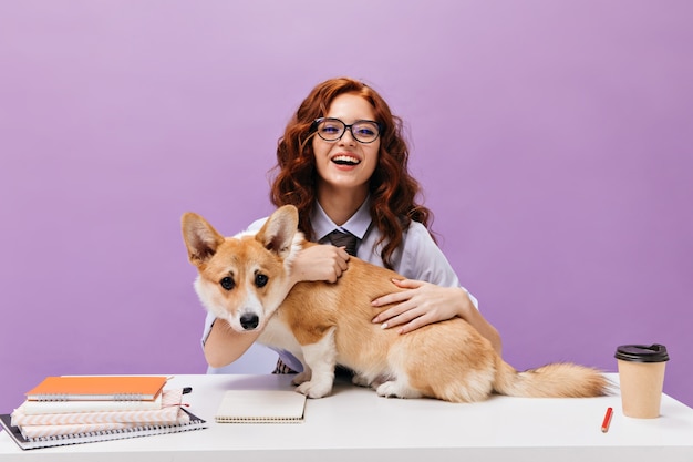 Curly lady in shirt and eyeglasses smiling and playing with corgi