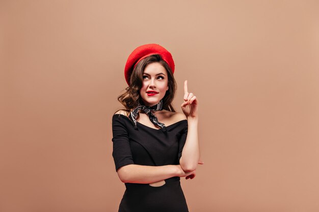 Curly lady with red lips looking up thoughtfully and showing index finger up. Portrait of woman in bright beret and black dress.