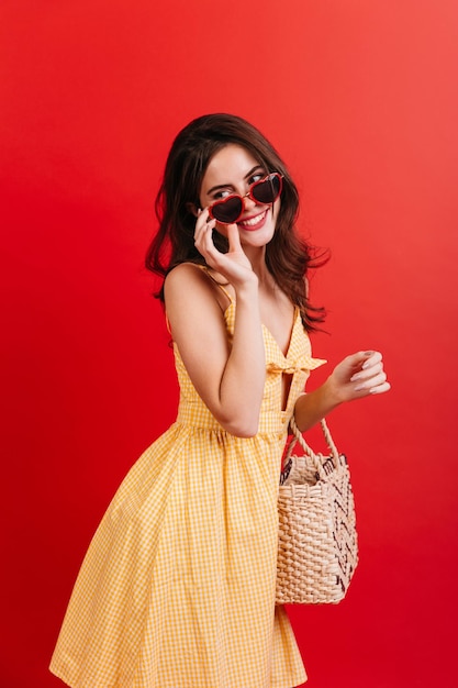 Curly lady in yellow dress in pinup style is smiling affably brunette with straw bag puts on sunglasses