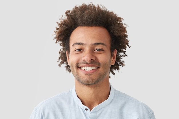 Curly man with broad smile, shows perfect teeth, being amused by interesting talk, has bushy curly dark hair stands indoor against white blank wall