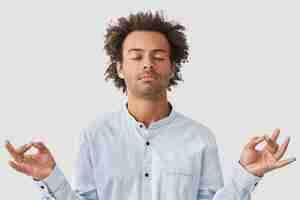 Free photo curly man with dark skin and curly hair, dressed in shirt, keeps eyes closed, holds hands in mudra sign, enjoys peaceful atmosphere