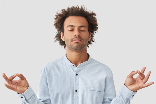 Curly man with dark skin and curly hair, dressed in shirt, keeps eyes closed, holds hands in mudra sign, enjoys peaceful atmosphere