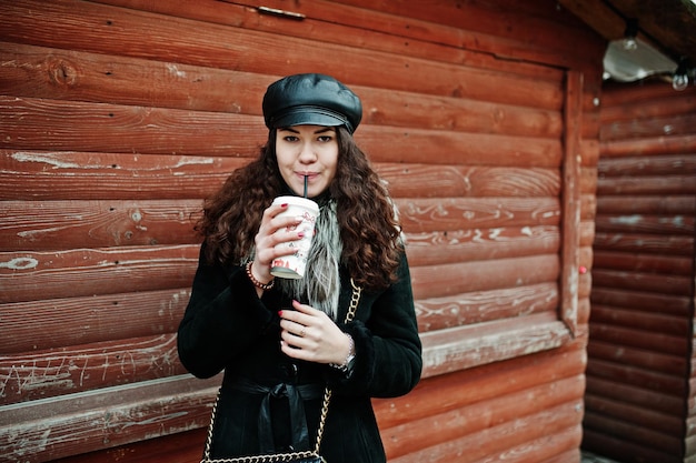 Curly mexican girl in leather cap and plastic cup of coffee at hand against wooden market