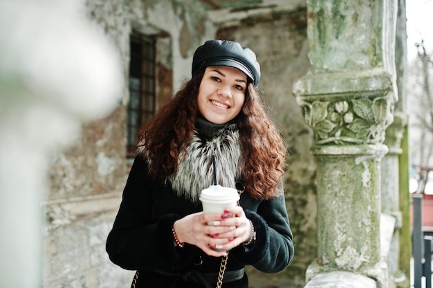 街の通りを歩いて手元に革のキャップとコーヒーのプラスチックカップで巻き毛のメキシコの女の子