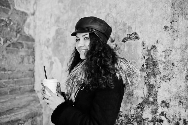 Curly mexican girl in leather cap and plastic cup of coffee at hand walking at streets of city