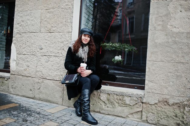 Curly mexican girl in leather cap and plastic cup of coffee at hand walking at streets of city