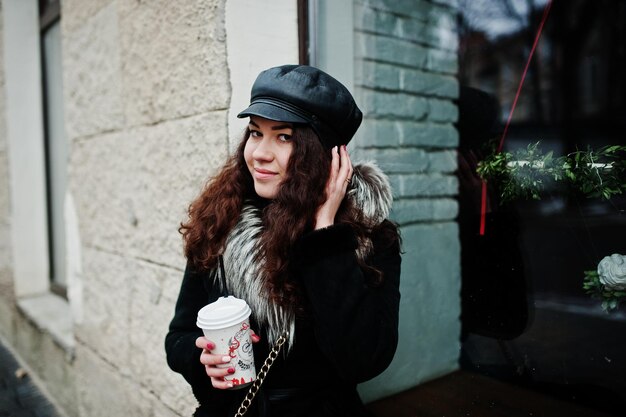 Curly mexican girl in leather cap and plastic cup of coffee at hand walking at streets of city