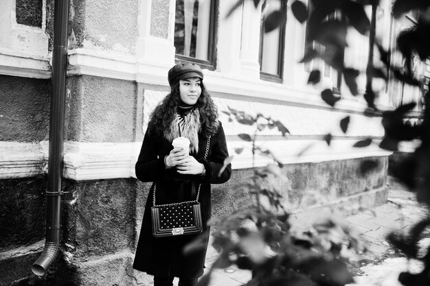 Curly mexican girl in leather cap and plastic cup of coffee at hand walking at streets of city